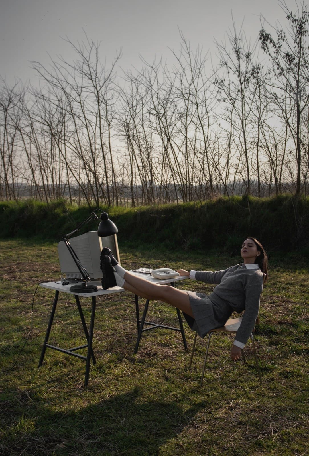 A woman sits at a small outdoor desk in a rural landscape, papers and laptop in front of her.