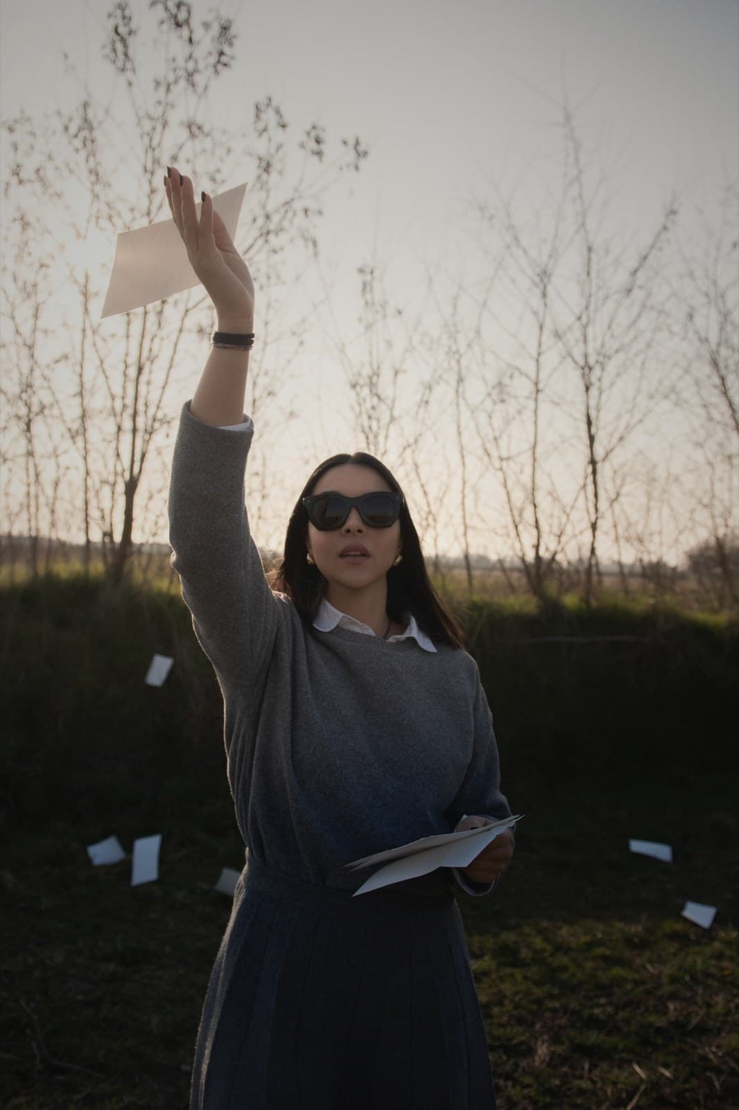 A woman in a coat releases a sheaf of papers into the air, a bare tree landscape behind her at dusk.