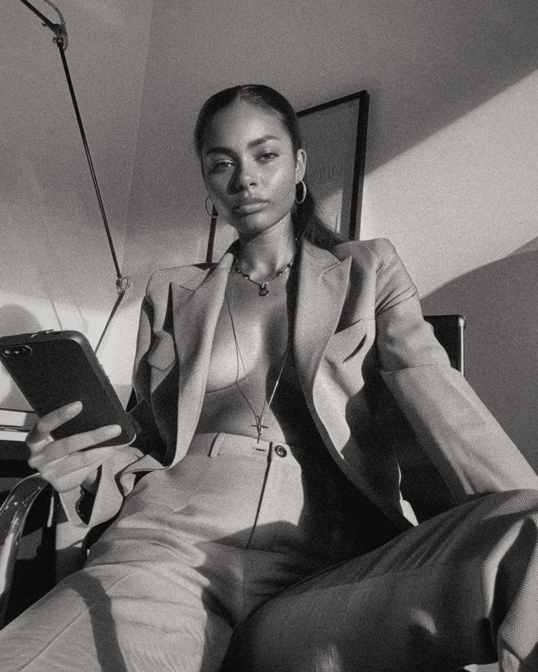 A woman in a tailored blazer sits back with her phone in hand, in warm directional light.