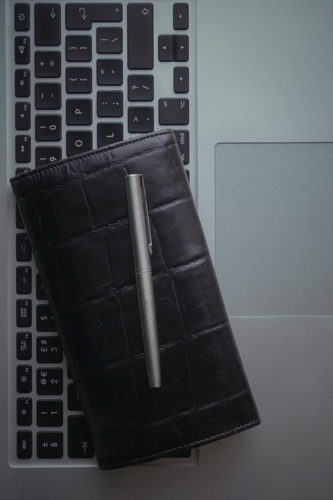 An overhead flat lay: a black leather wallet and a pen resting across a laptop keyboard.
