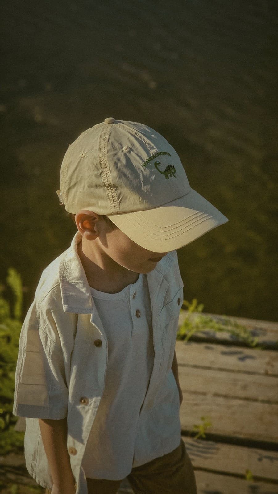 A child in a cream outfit crouches at the water's edge in soft light, sleeves rolled, deep in a quiet moment with the tide.