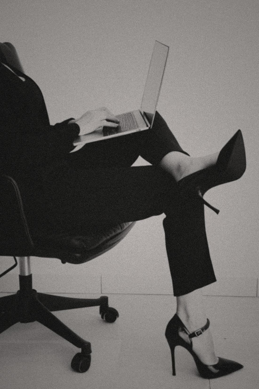 A woman in a sharp suit and pointed heels reclines in an office chair, laptop balanced on her lap.