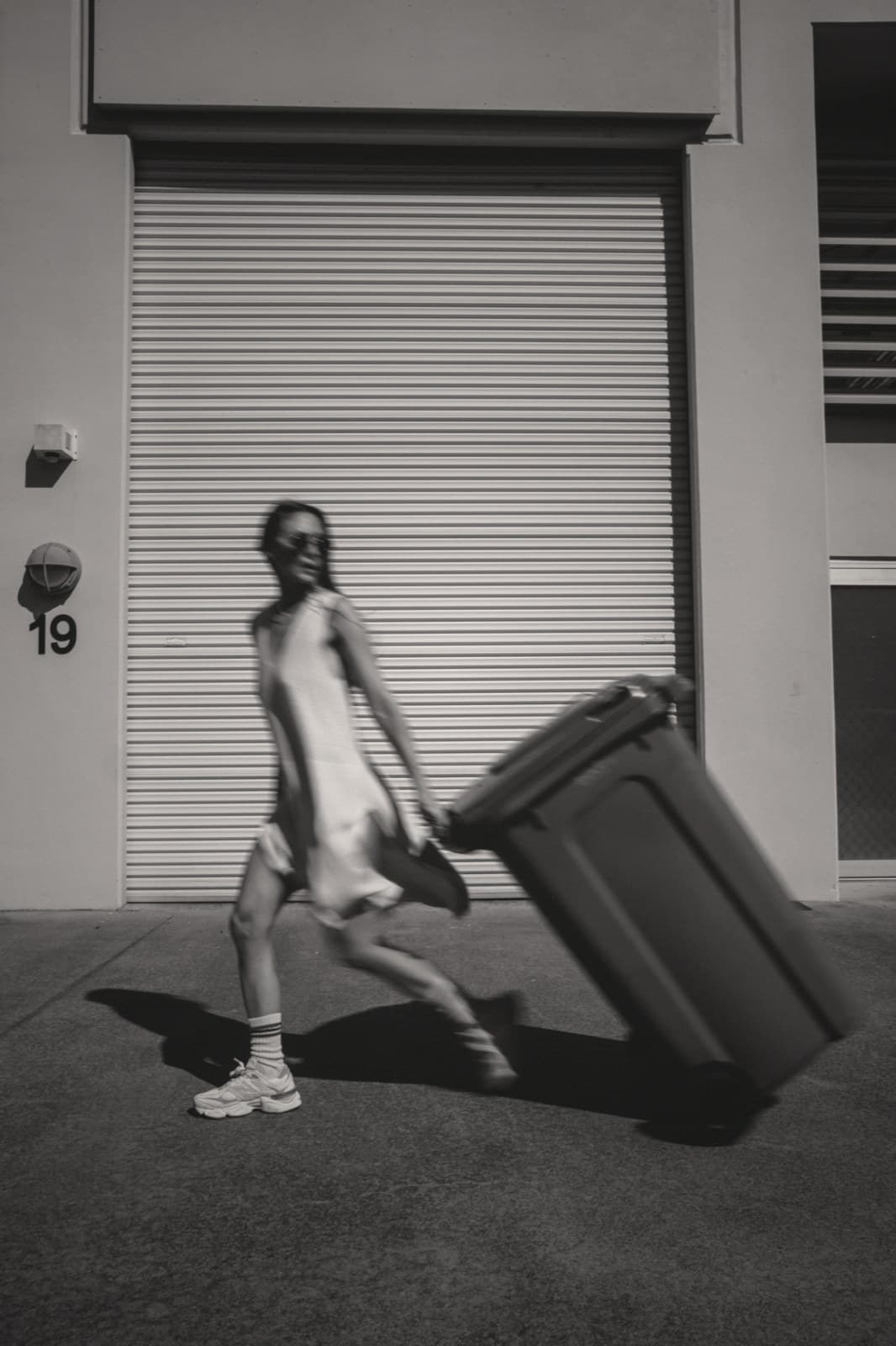 A woman walks an oversized trash bin down an urban sidewalk, mid-stride, midday light.