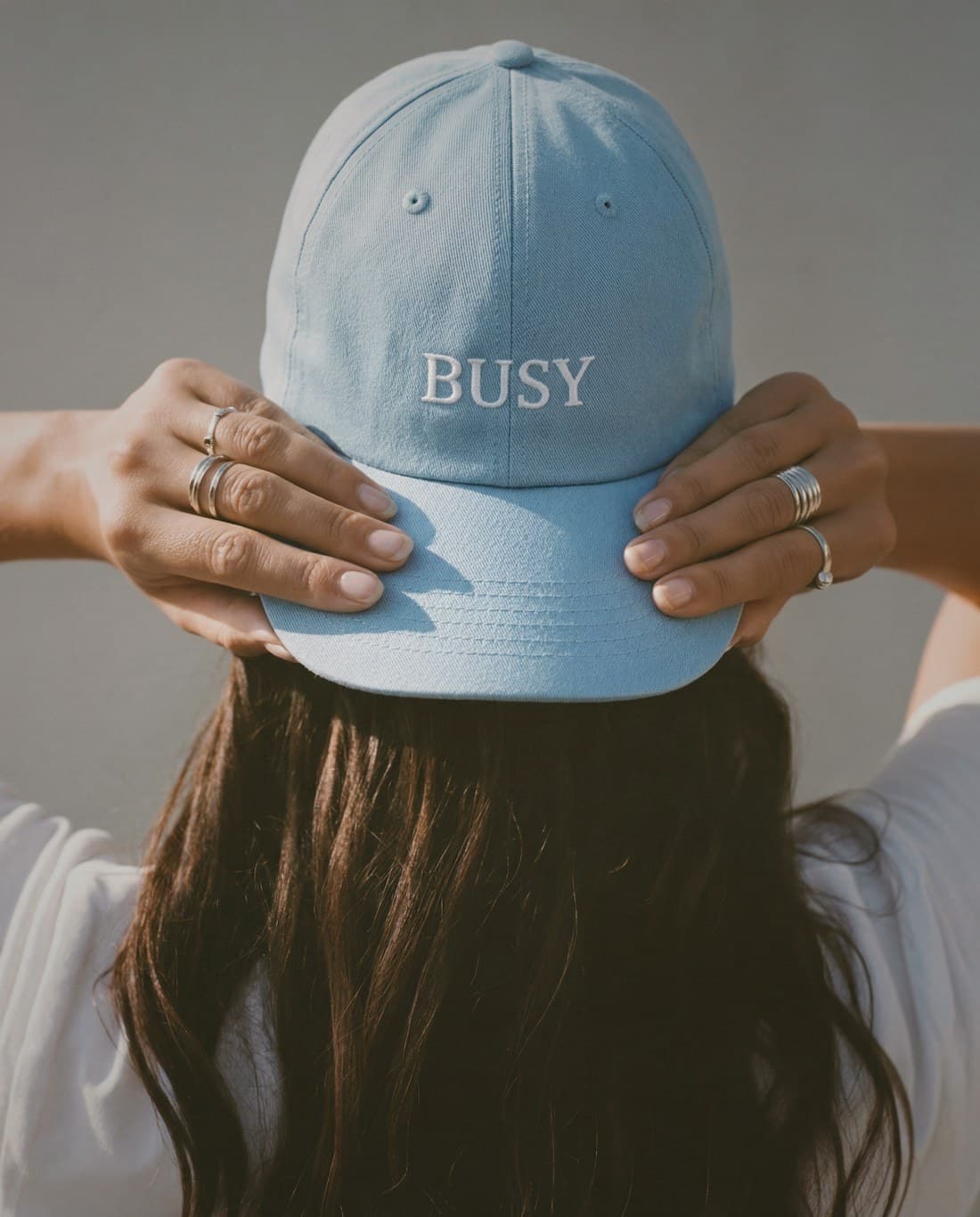 A woman seen from behind holds a pale blue cap reading BUSY above her head.