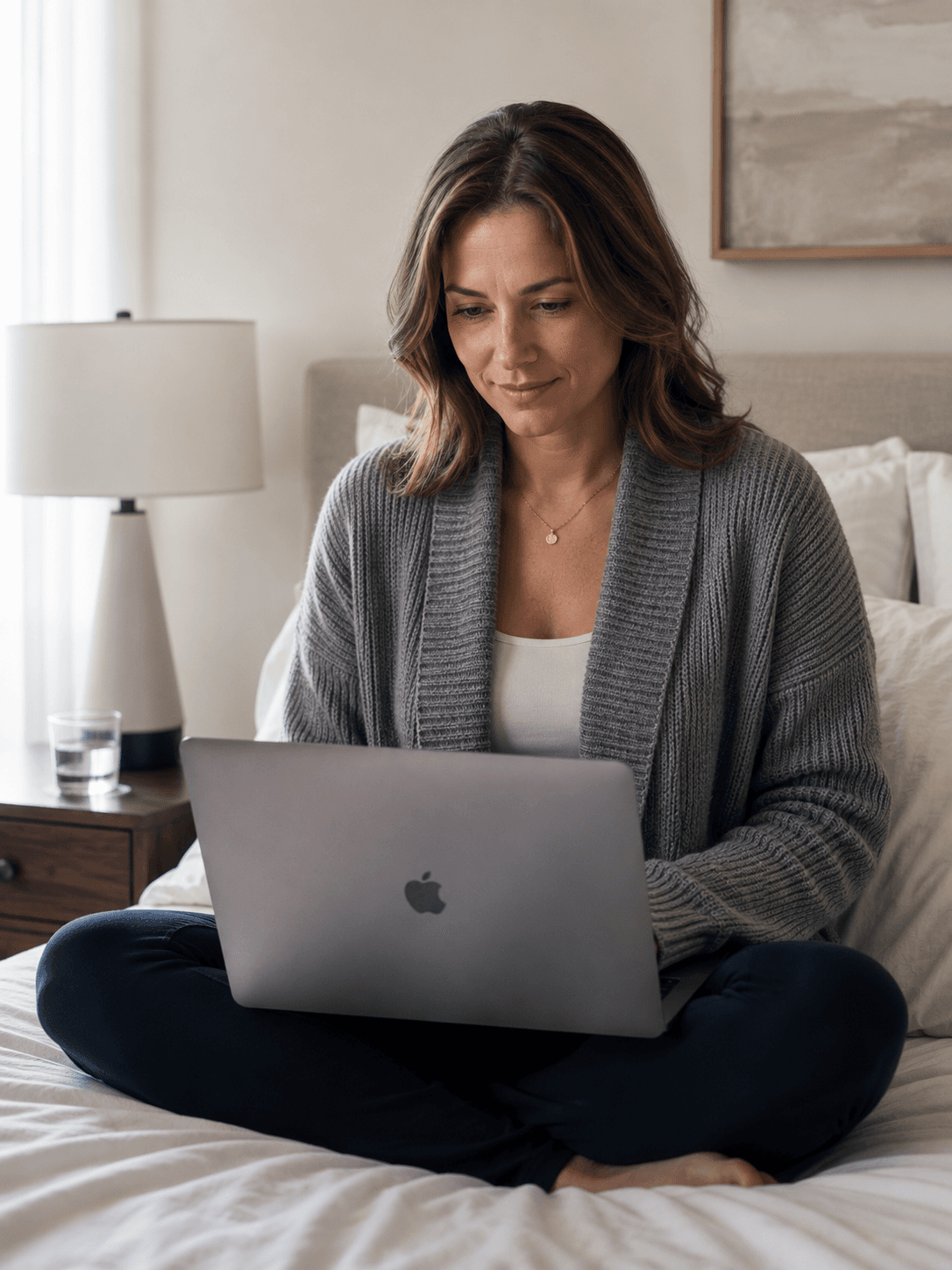 A mother in a gray cardigan sits cross-legged on her bed with a laptop, the lamp on, the day finally still.
