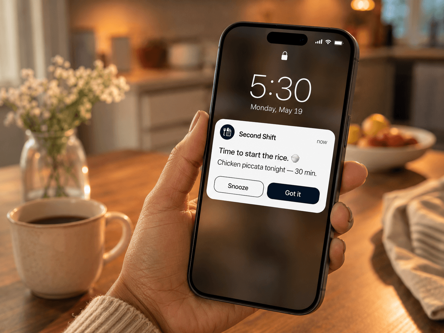 A hand holding a phone in a warm kitchen at 5:30. A Second Shift notification reads Time to start the rice, chicken piccata tonight, 30 minutes, with Snooze and Got it buttons.